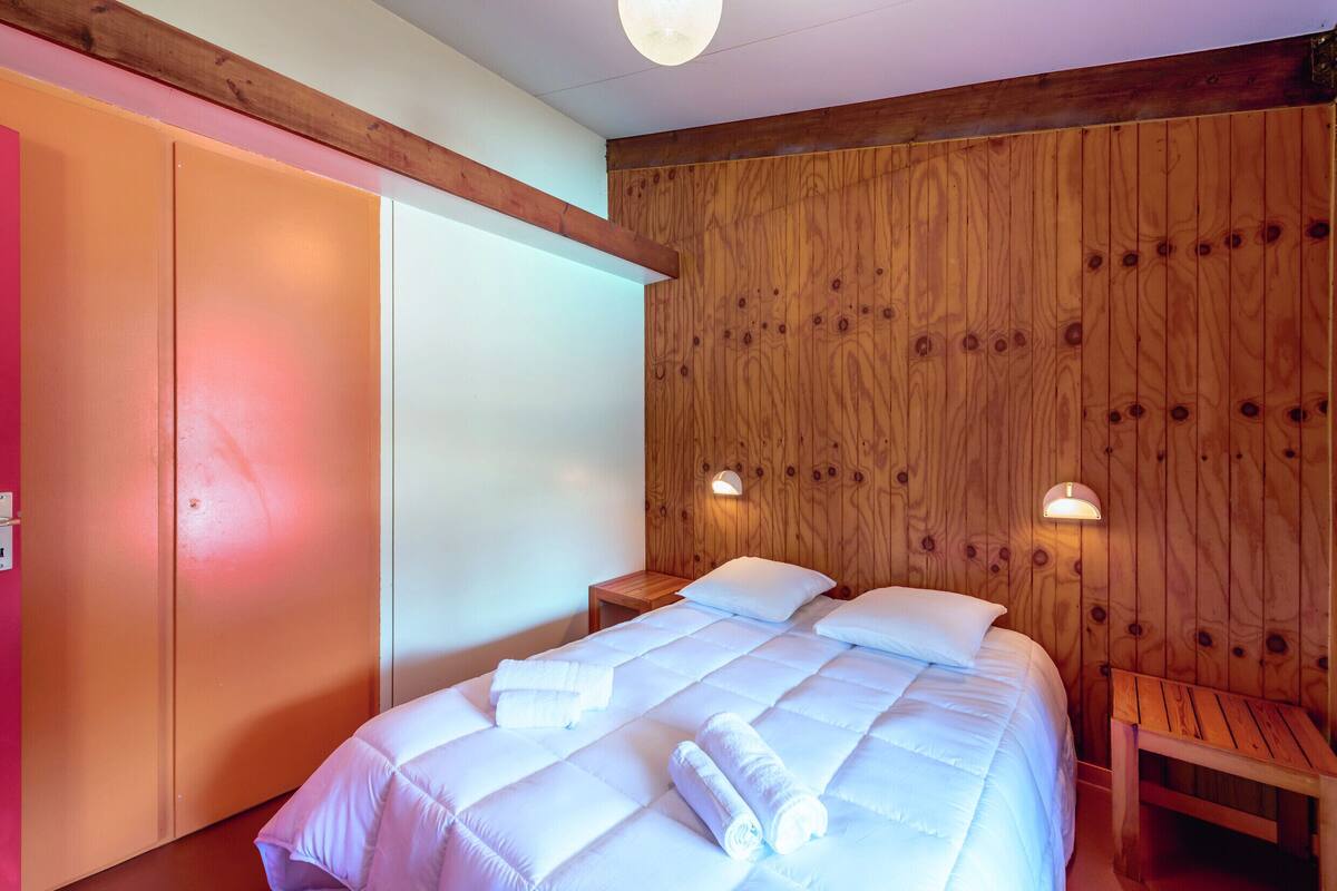 Un dormitorio con cabecera de madera, cama con ropa de cama blanca, dos lámparas de noche y un banco de madera.