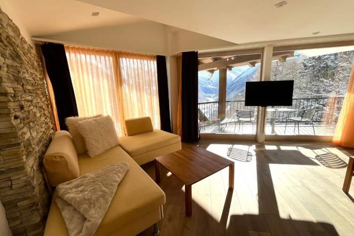 A living room with a sofa, a coffee table, a TV, and a balcony with a view of mountains.