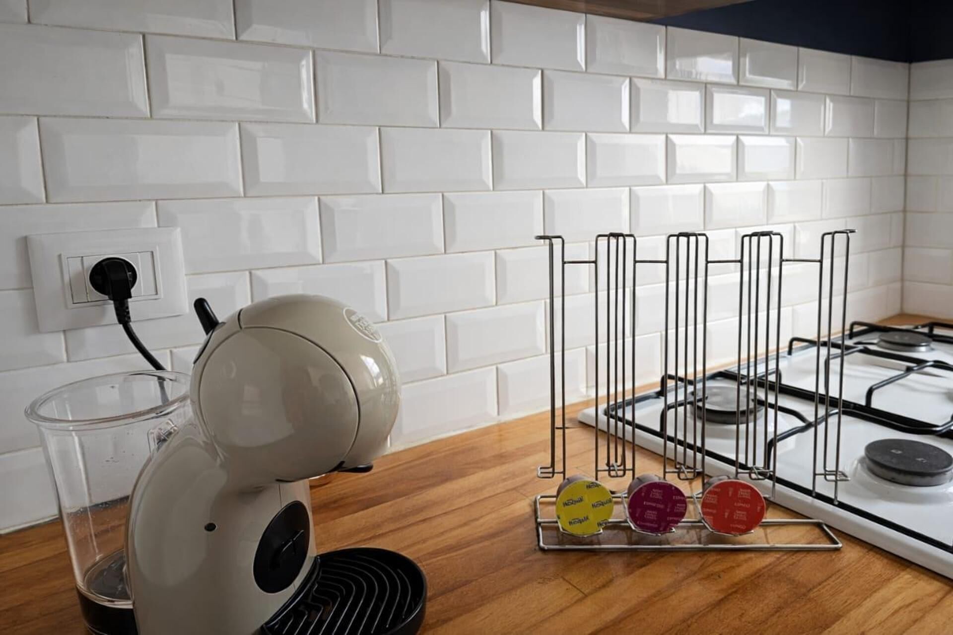 A kitchen with white tiles, a gas stove, a kettle, and a kettle holder with tea bags.