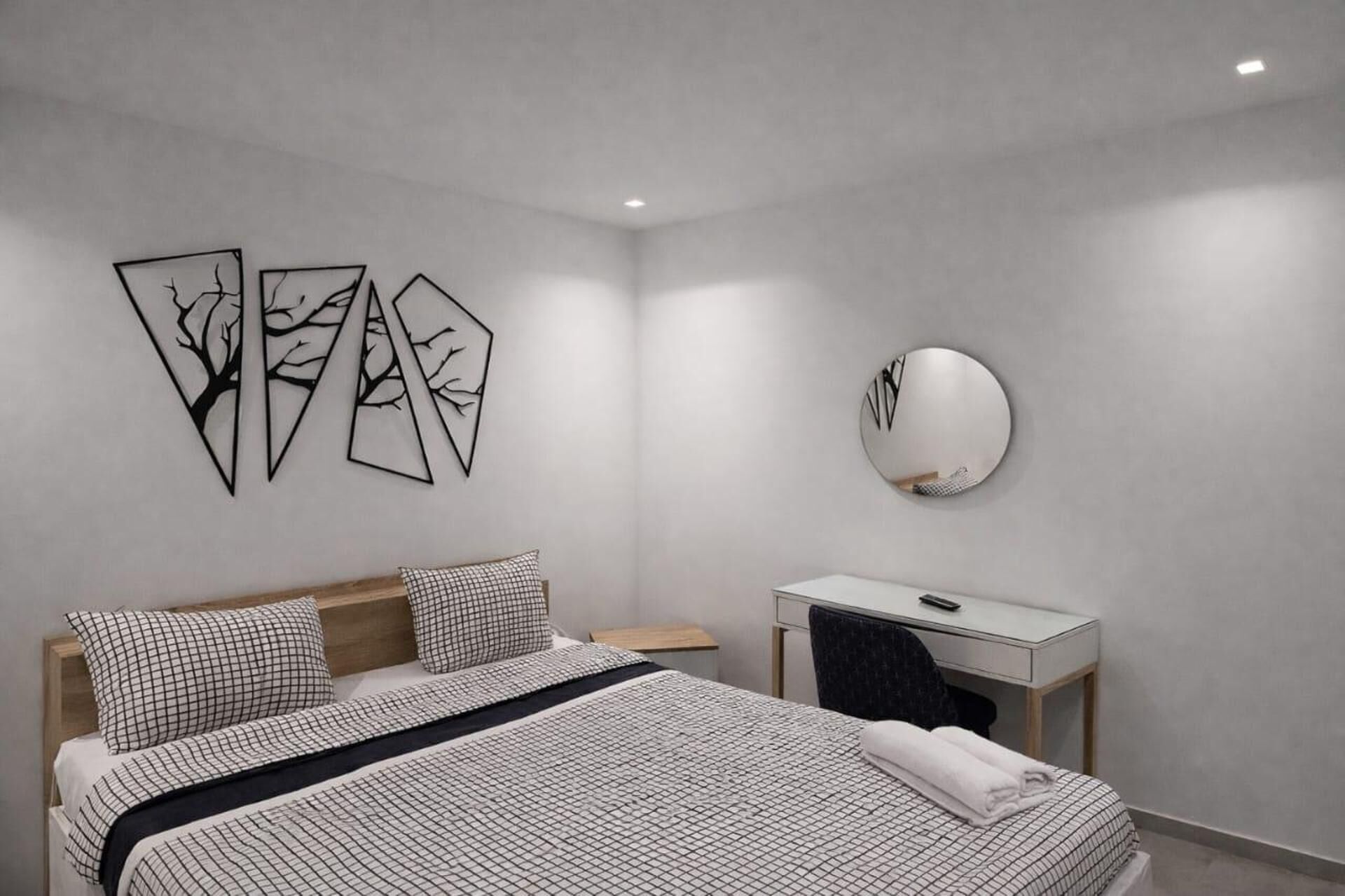 Quarto moderno com cama, mesa de cabeceira e escrivaninha. Há obras de arte abstratas nas paredes e um espelho redondo.