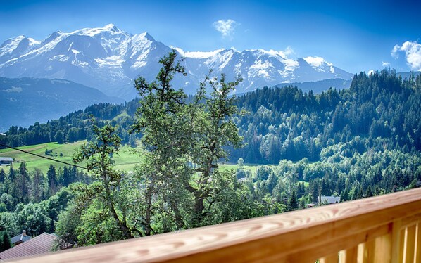 A mountain landscape with snow-capped peaks, dense forest, and a wooden balcony railing.