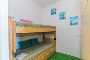 A bunk bed with a green patterned bedspread and two pillows, a wooden headboard, and a small window with a view of the sea.