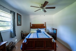 Un dormitorio con una cama de madera, un ventilador de techo, una mesita de noche con lámpara, una silla y una ventana con cortinas.
