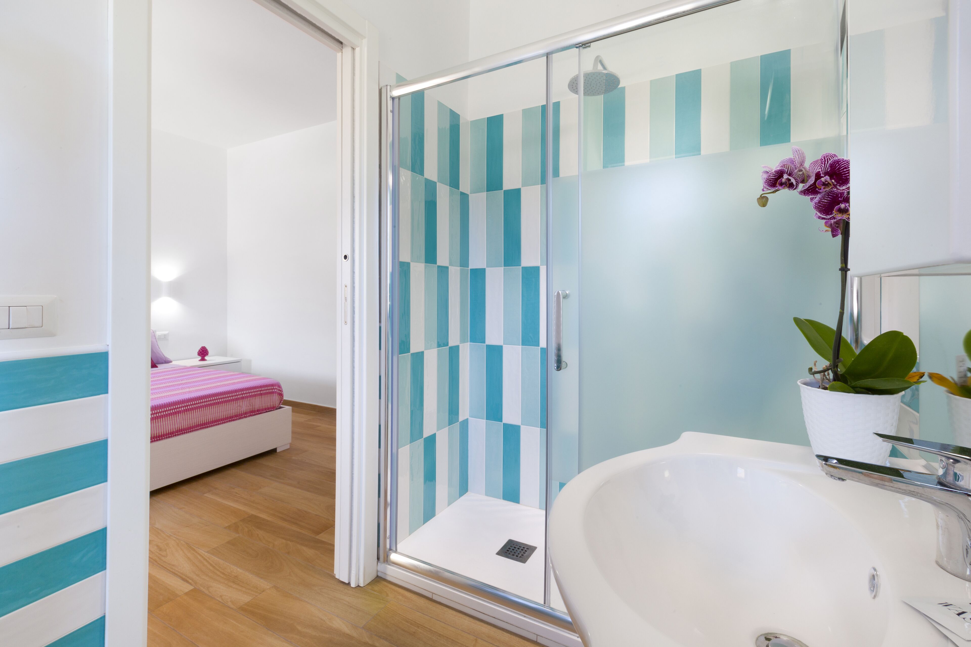 A modern bathroom with a glass shower enclosure, a white sink, and a bed with a striped bedspread.