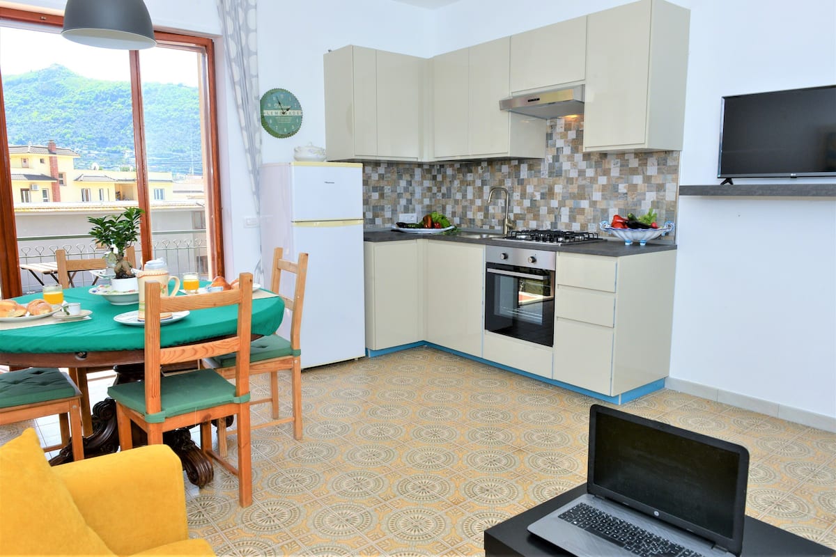 Cocina compacta con comedor, una laptop sobre una mesa, y vista de edificios a través de una ventana.
