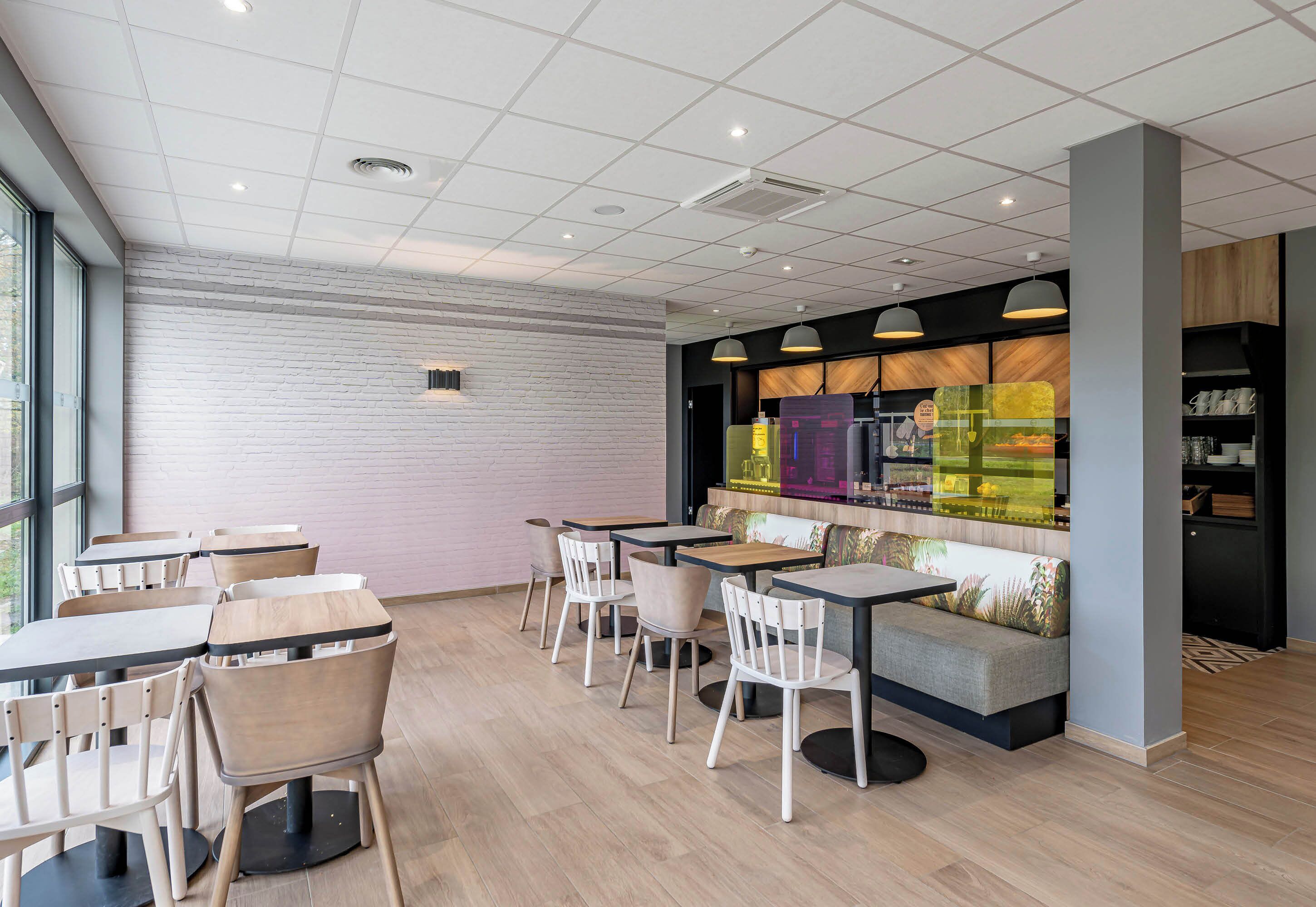 A modern cafe with wooden tables and chairs, a seating area with cushions, and a large window providing natural light.