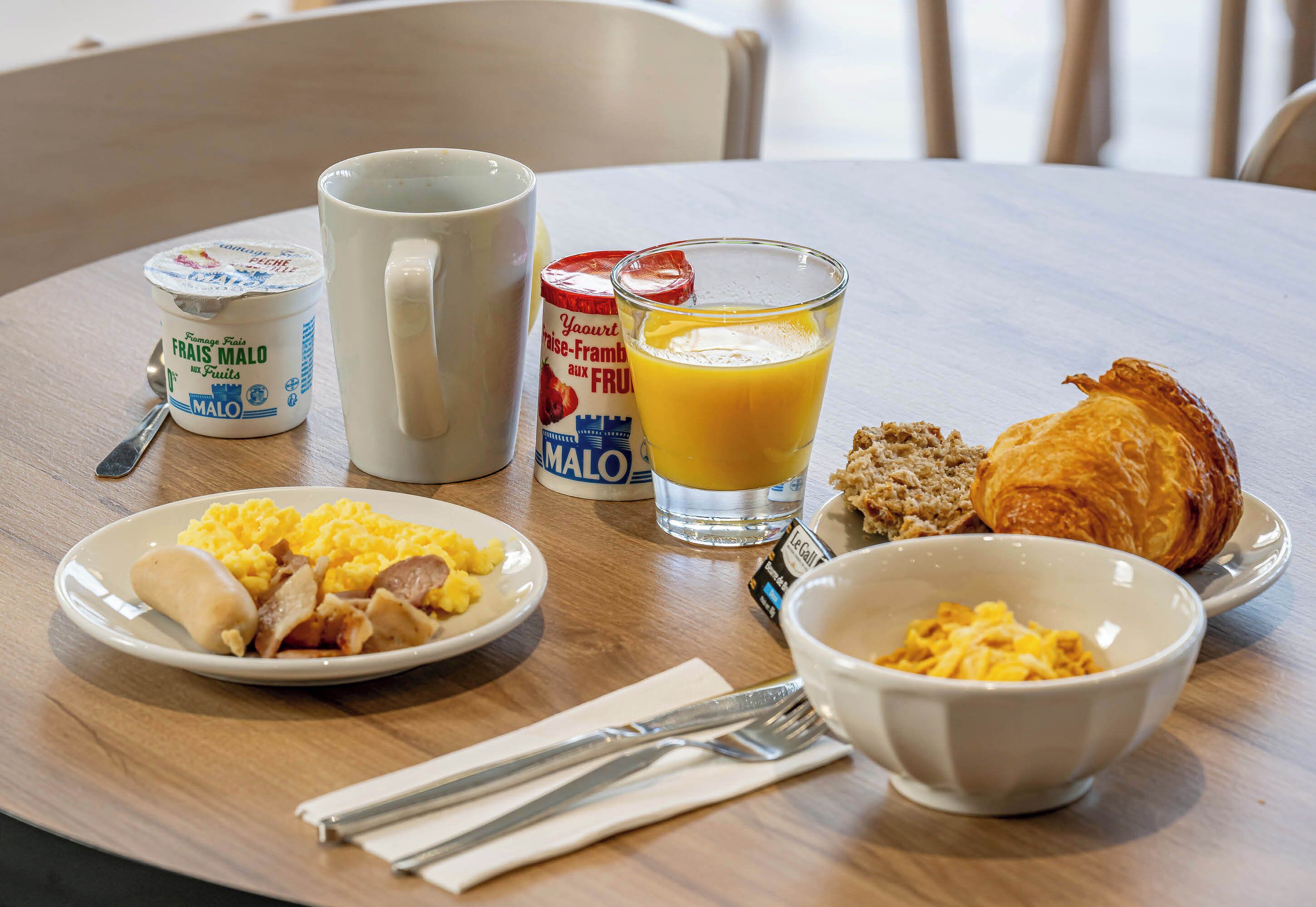 Una tavola imbandita per la colazione con uova strapazzate, salsiccia, pane tostato, un bicchiere di succo d'arancia, una ciotola di cereali e un croissant.