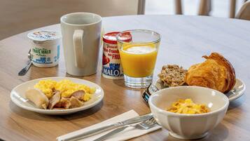 Una tavola imbandita per la colazione con uova strapazzate, salsiccia, pane tostato, un bicchiere di succo d'arancia, una ciotola di cereali e un croissant.