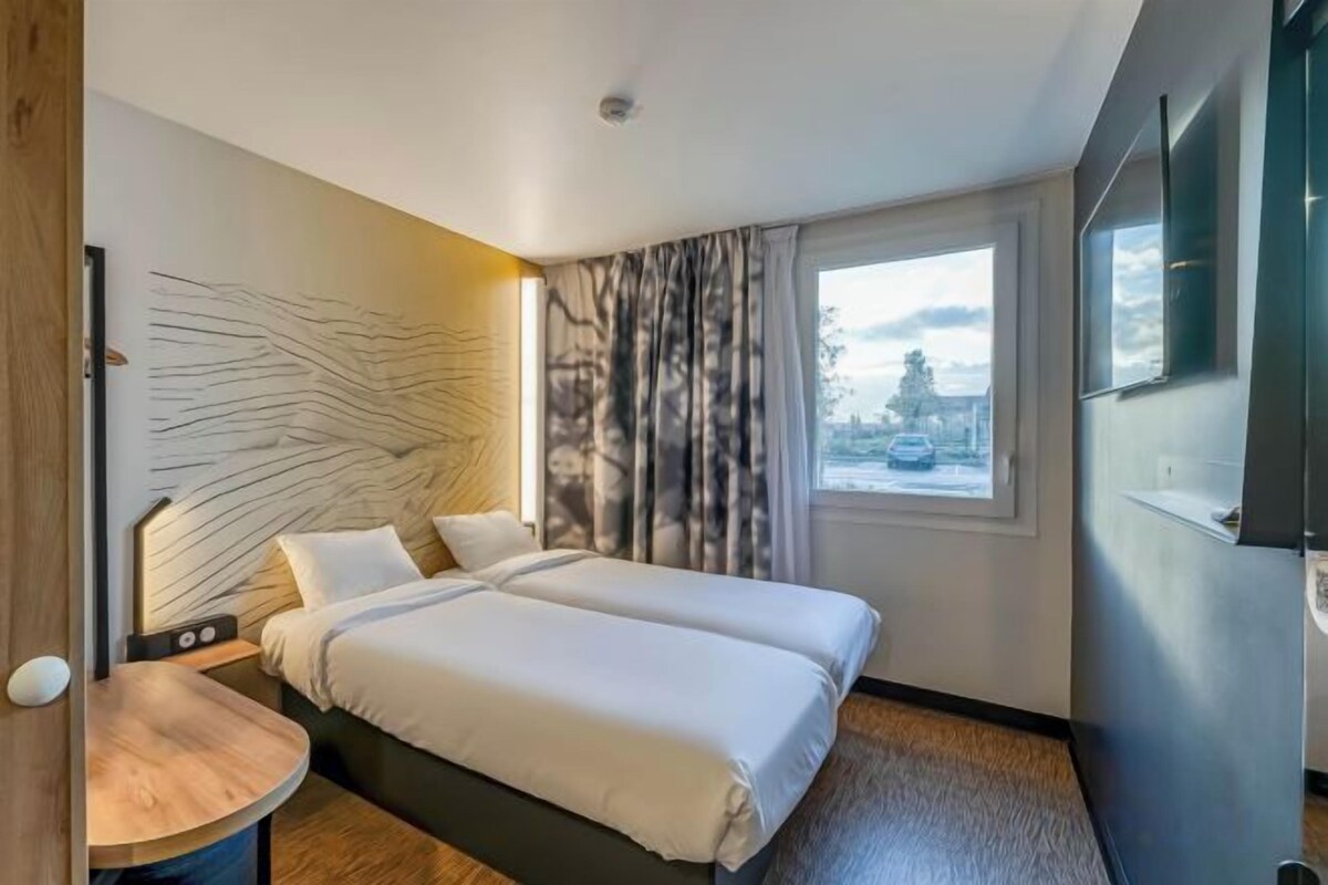 Ett hotellrum med två sängar, ett träbord, ett stort fönster med gardiner och utsikt över omgivningen.
