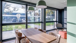 A modern conference room with a large table and chairs, large windows with a view of a parking lot and trees, and pendant lighting. - B&B HOTEL Bordeaux Saint André De Cubzac (Saint-André-de-Cubzac)