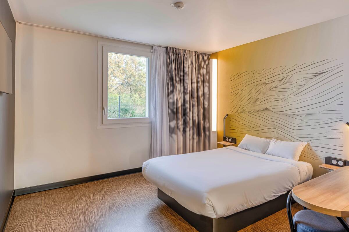 Ein modernes Hotelzimmer mit einem großen Bett, einem kleinen Tisch und einem Fenster mit Vorhängen.