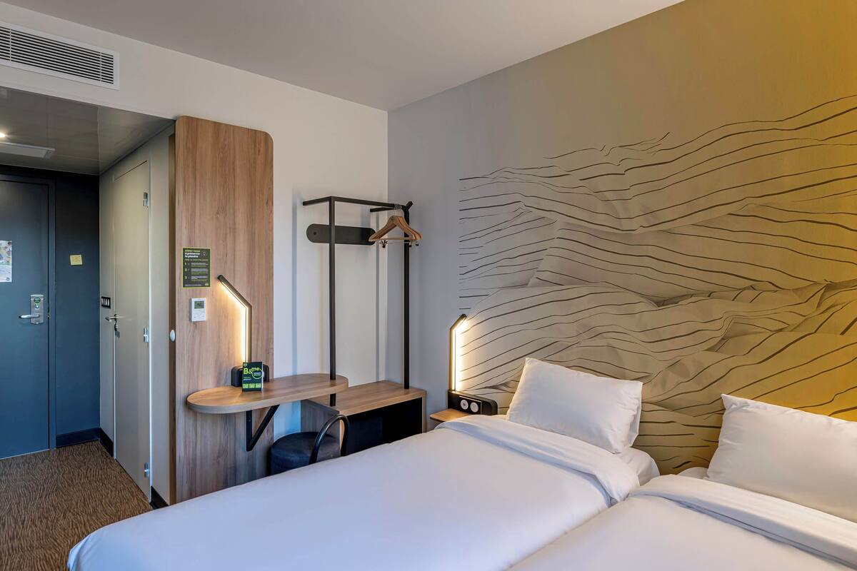 Ein Hotelzimmer mit Bett, Schreibtisch mit Stuhl, Kleiderständer, Wanduhr und Tür.