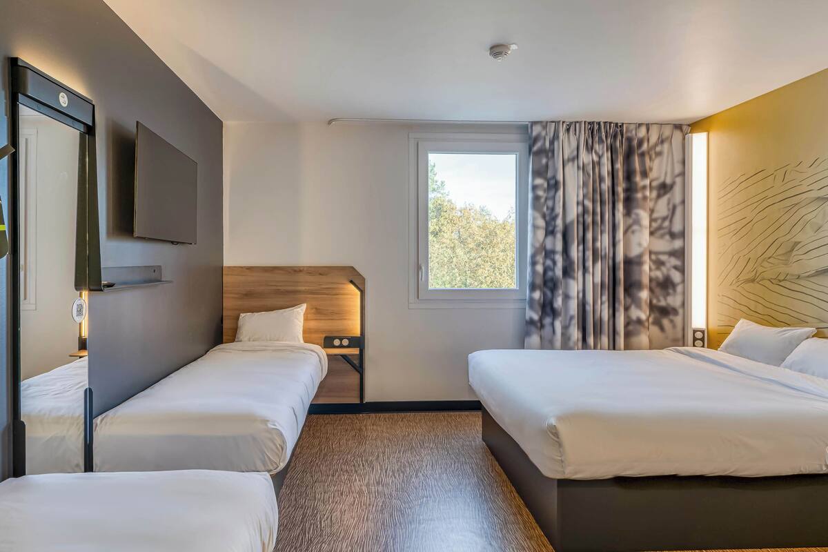 Ein Hotelzimmer mit zwei Einzelbetten, einem Holzkopfteil, einem Flachbildfernseher und einem Fenster mit Vorhängen.
