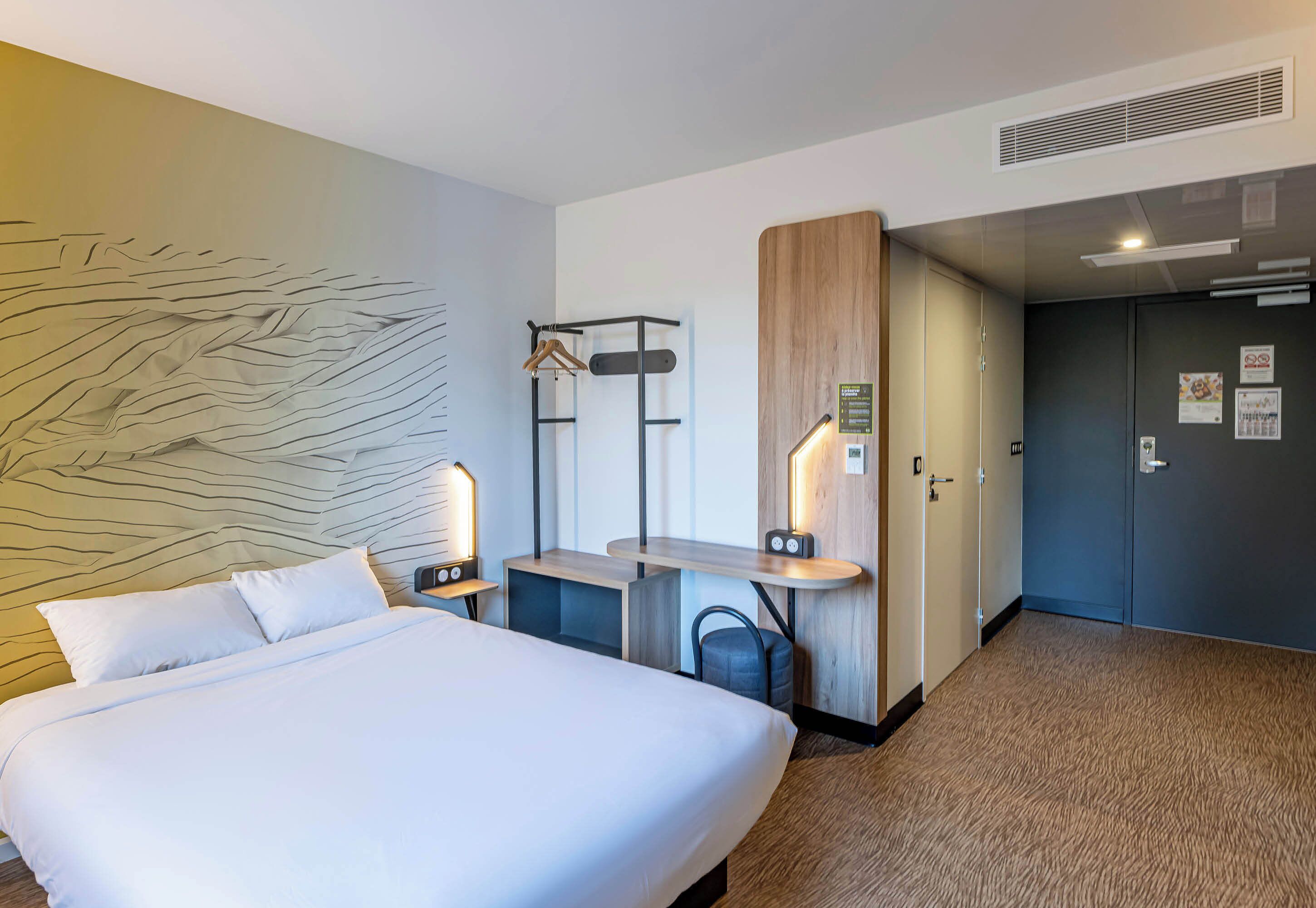 A hotel room with a large bed, a desk with a chair, a coat rack, and a wardrobe.