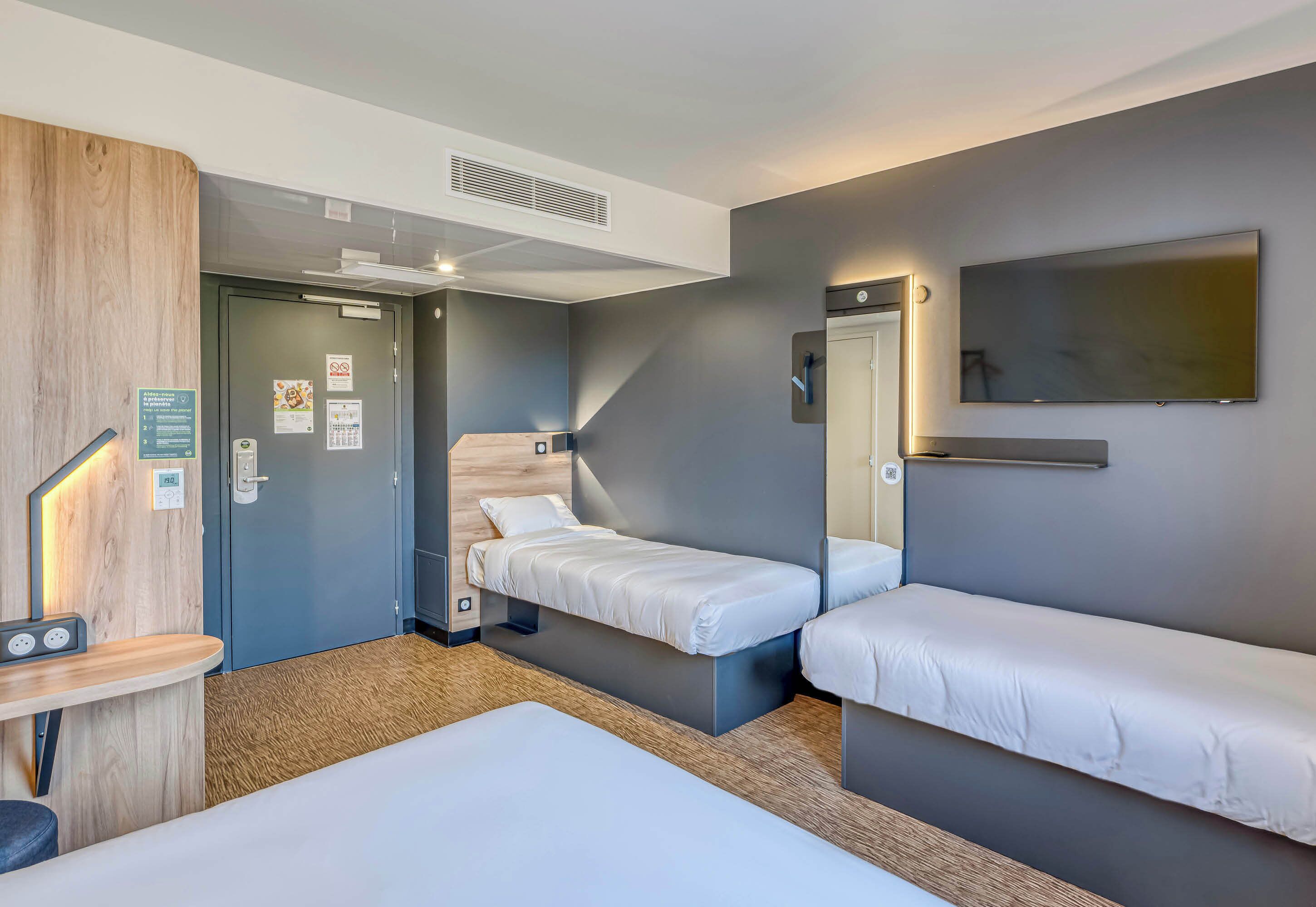 A hotel room with two single beds, a desk with a chair, a small table, a TV mounted on the wall, and a door leading to another room.