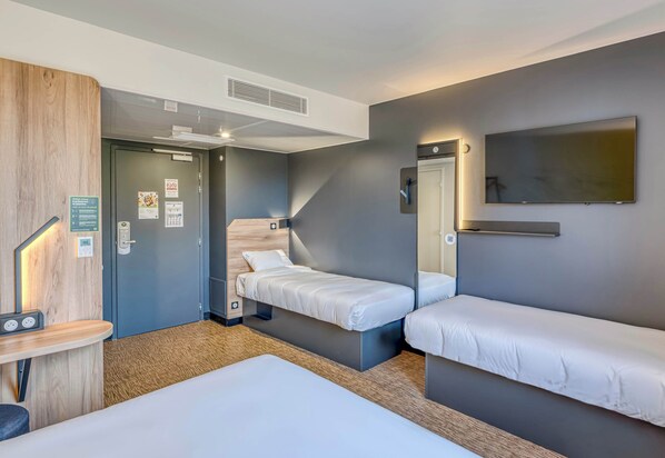 Habitación de hotel con dos camas individuales, un escritorio con silla, una mesita pequeña, un televisor montado en la pared y una puerta que da a otra habitación.