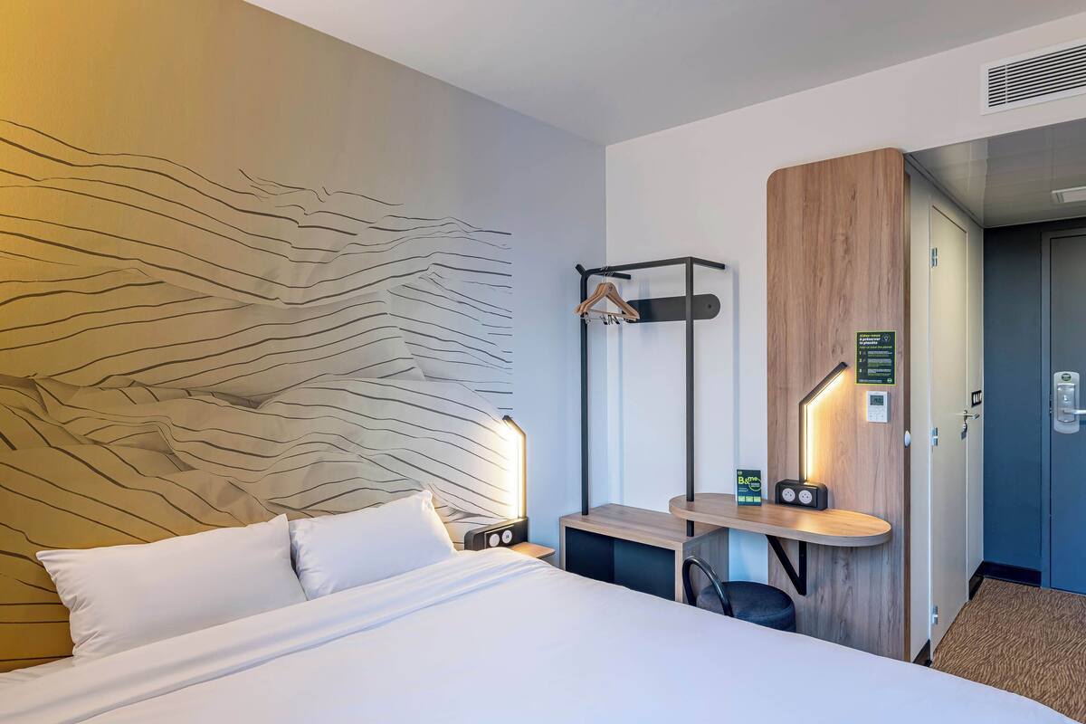 Ein Hotelzimmer mit Bett, Nachttischen, einem hölzernen Kopfteil, einem Kleiderhaken und einem Schreibtisch mit Stuhl.