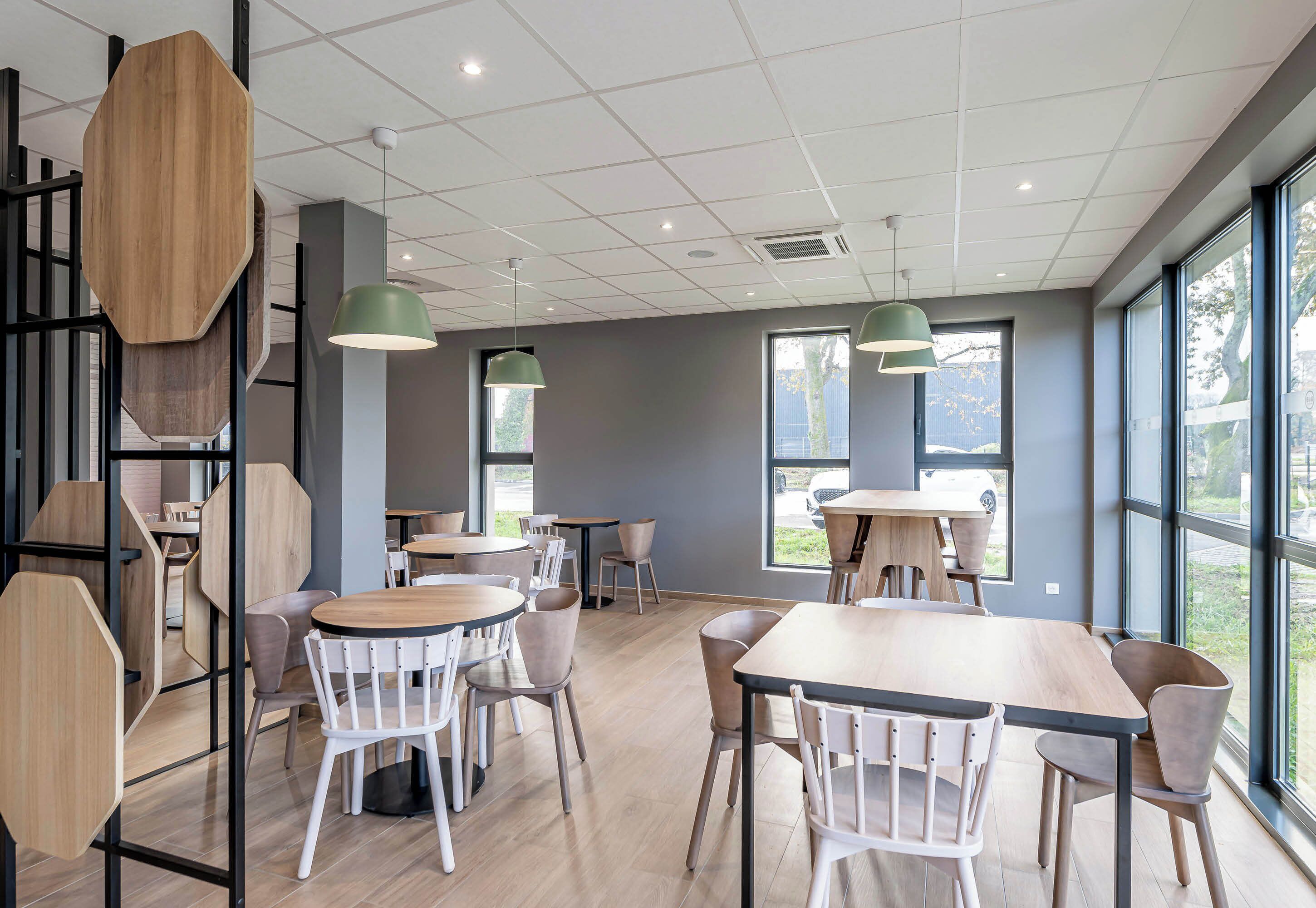 A modern cafe with wooden tables and chairs, large windows, and green pendant lights.