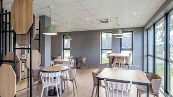 A modern cafe with wooden tables and chairs, large windows, and green pendant lights.