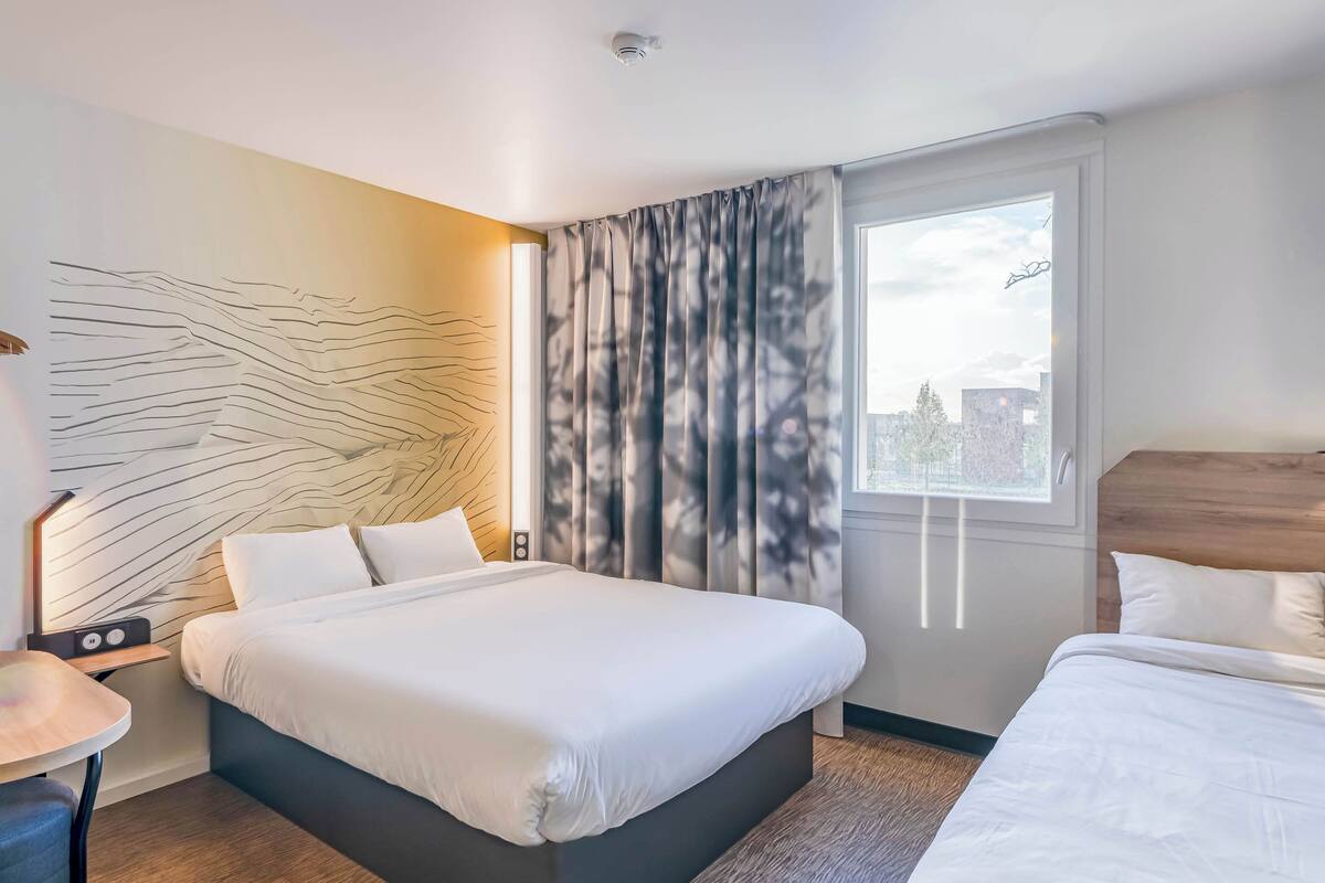 Ein modernes Hotelzimmer mit einem großen Bett, einem Nachttisch mit Lampe und Blick auf die Stadt durch ein Fenster mit Vorhängen.