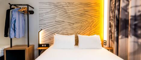 A modern hotel room with a large bed, a bedside table with a lamp, a wardrobe, and a wall with a unique textured design.