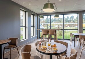 A modern dining area with round tables and chairs, large windows offering a view of the outdoors, and pendant lighting. - B&B HOTEL Bordeaux Saint André De Cubzac (Saint-André-de-Cubzac)