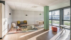 A modern office space with a meeting area, a sofa, a table, and a large window providing a view of the outside. - B&B HOTEL Bordeaux Saint André De Cubzac (Saint-André-de-Cubzac)