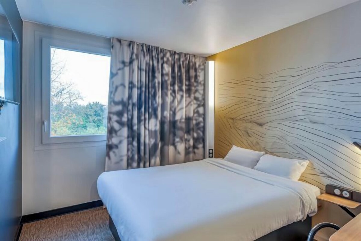 A modern hotel room with a large bed, a window with curtains, a headboard with a unique design, and a bedside table with a lamp.