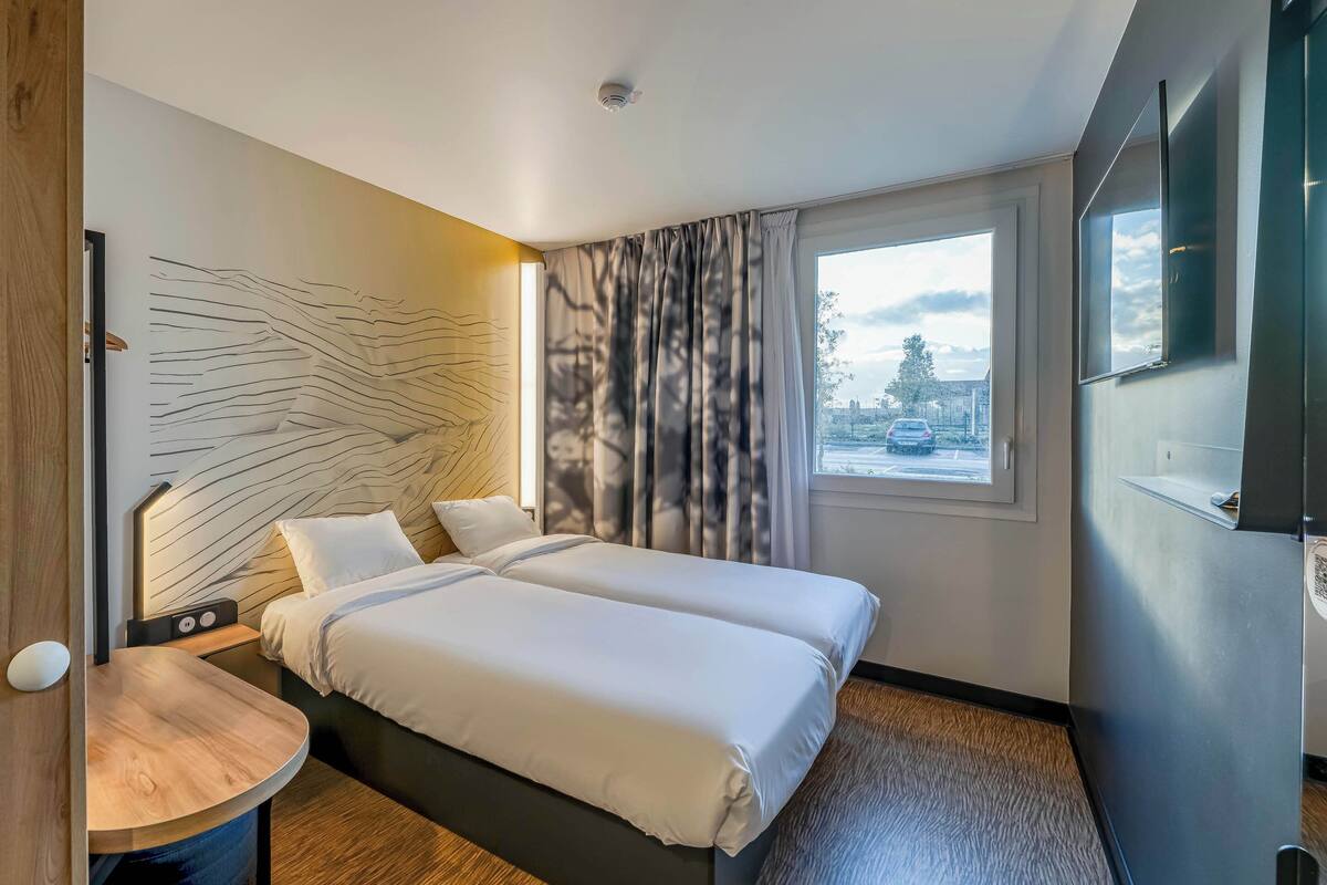 Ein modernes Hotelzimmer mit Doppelbett, Nachttisch, Wand-TV und einem Fenster mit Vorhängen.