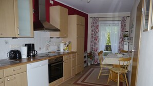 A compact kitchen with basic appliances, a dining area with a table and chairs, and a window with floral curtains. - Apartment on the 1st floor. Only 10 minutes walk to the town center. (Berchtesgaden)