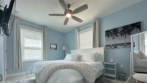 Un dormitorio con cama, ventilador de techo, mesita de noche y espejo, y ventana con persianas.