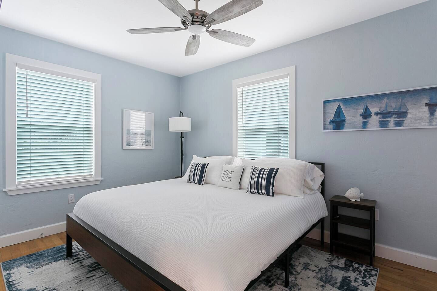 A bedroom with a bed, a ceiling fan, two windows with blinds, a nightstand, a lamp, and a painting on the wall.