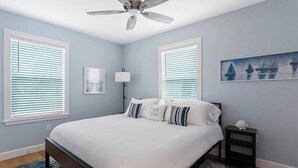 A bedroom with a bed, a ceiling fan, two windows with blinds, a nightstand, a lamp, and a painting on the wall.