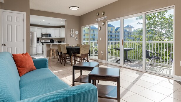 A spacious living area with a sofa, a dining table with chairs, a kitchen in the background, and a balcony with a view of buildings and greenery. - Princess Irene Unit 202 Oceanside 3BR 2BathCondo +Jacuzzi and parking (Ocean City)