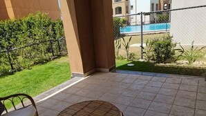 En uteplats med ett runt bord och flätade stolar, en liten trädgård med gräsmatta och buskar, och en utsikt över en swimmingpool och byggnader i bakgrunden.