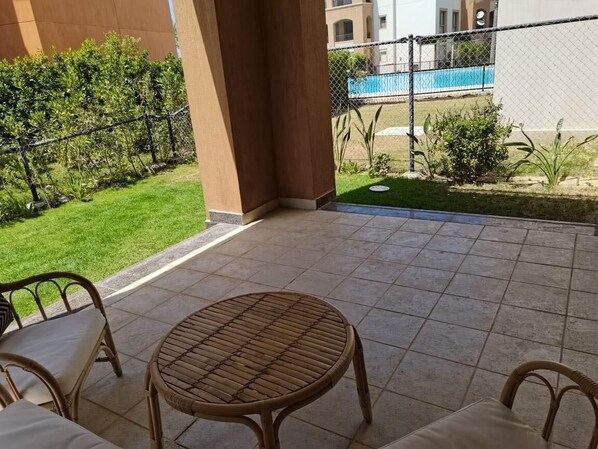 A patio area with a round table and wicker chairs, a small garden with a lawn and shrubs, and a view of a swimming pool and buildings in the background.
