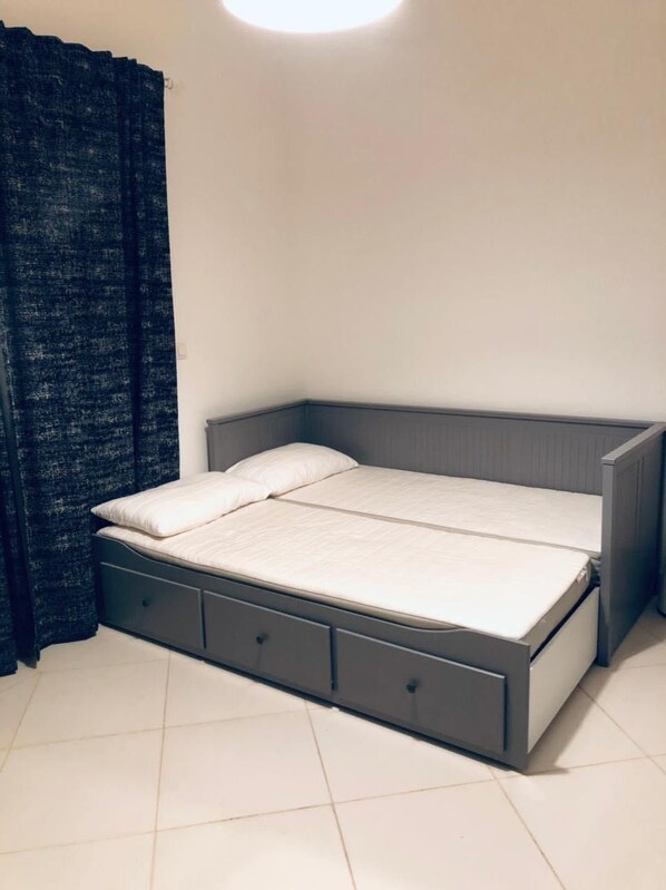 A single bed with a grey headboard and drawers, a dark curtain, and a white tiled floor.