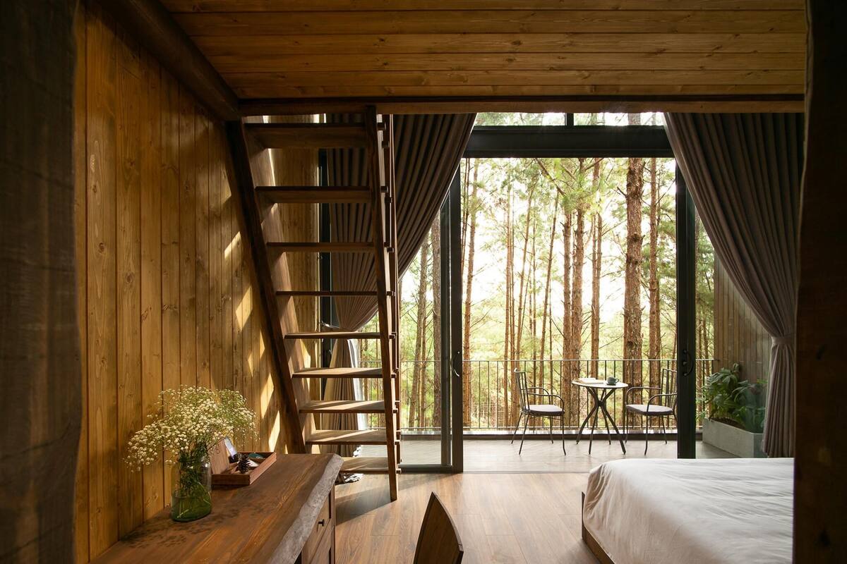 A wooden cabin room with a bed, a desk, a chair, a ladder, and a balcony with a table and chairs.