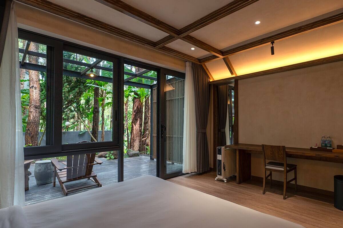 A hotel room with a large bed, a desk, and a view of a tropical garden through the glass door.