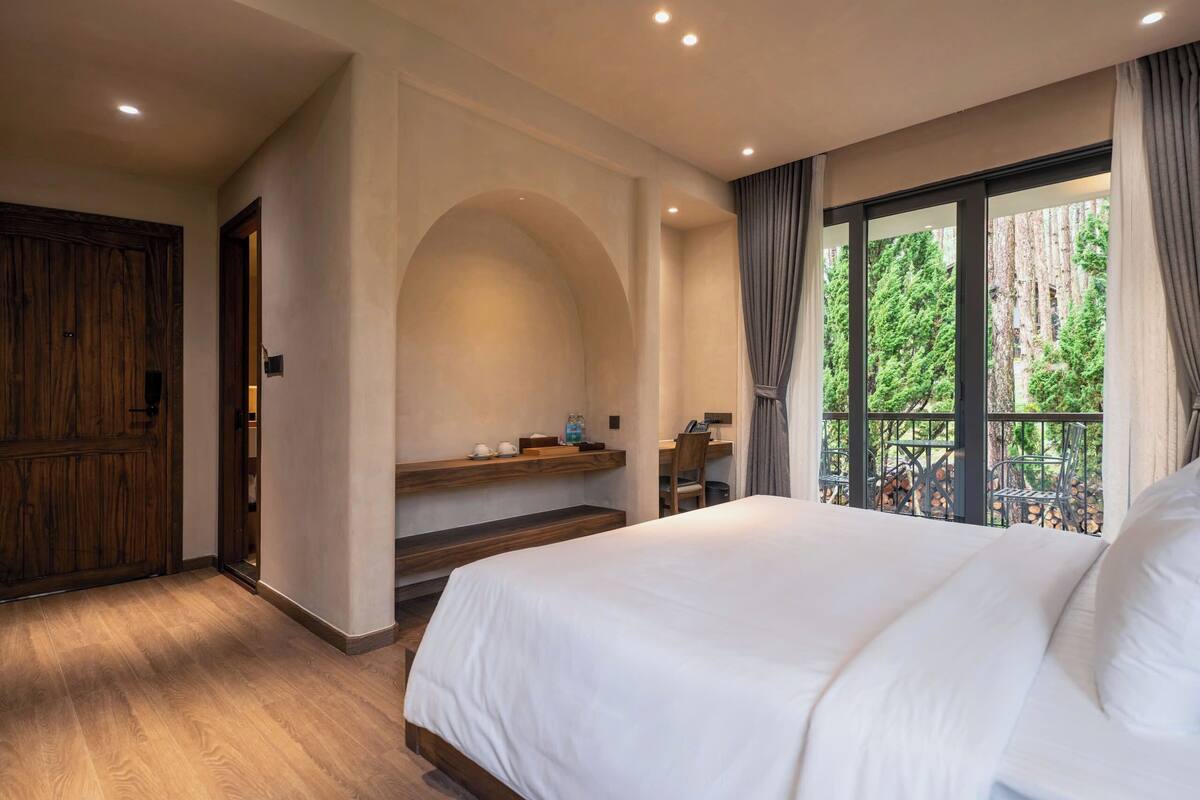 A bedroom with a large bed, a wooden door, a built-in bench, and a window with a view of trees.