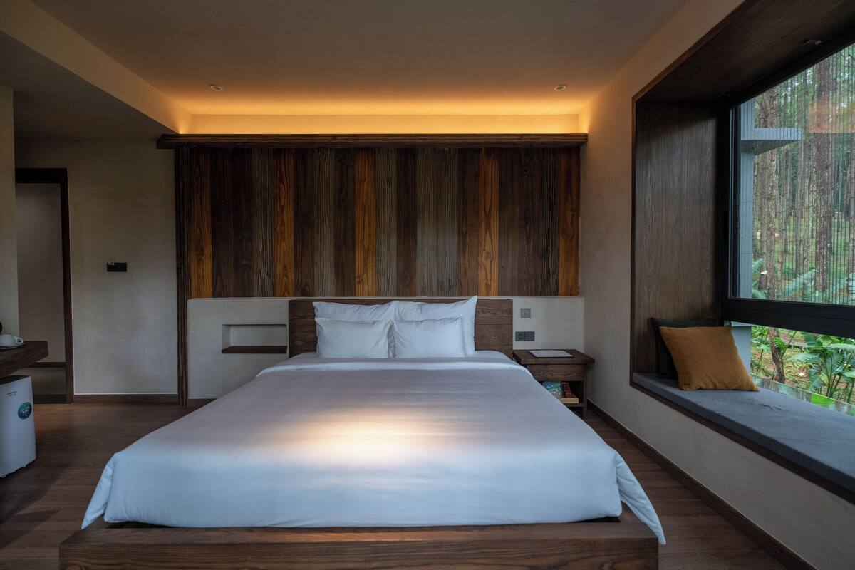 A modern bedroom with a large bed, wooden headboard, bedside tables, and a window with a view of greenery.