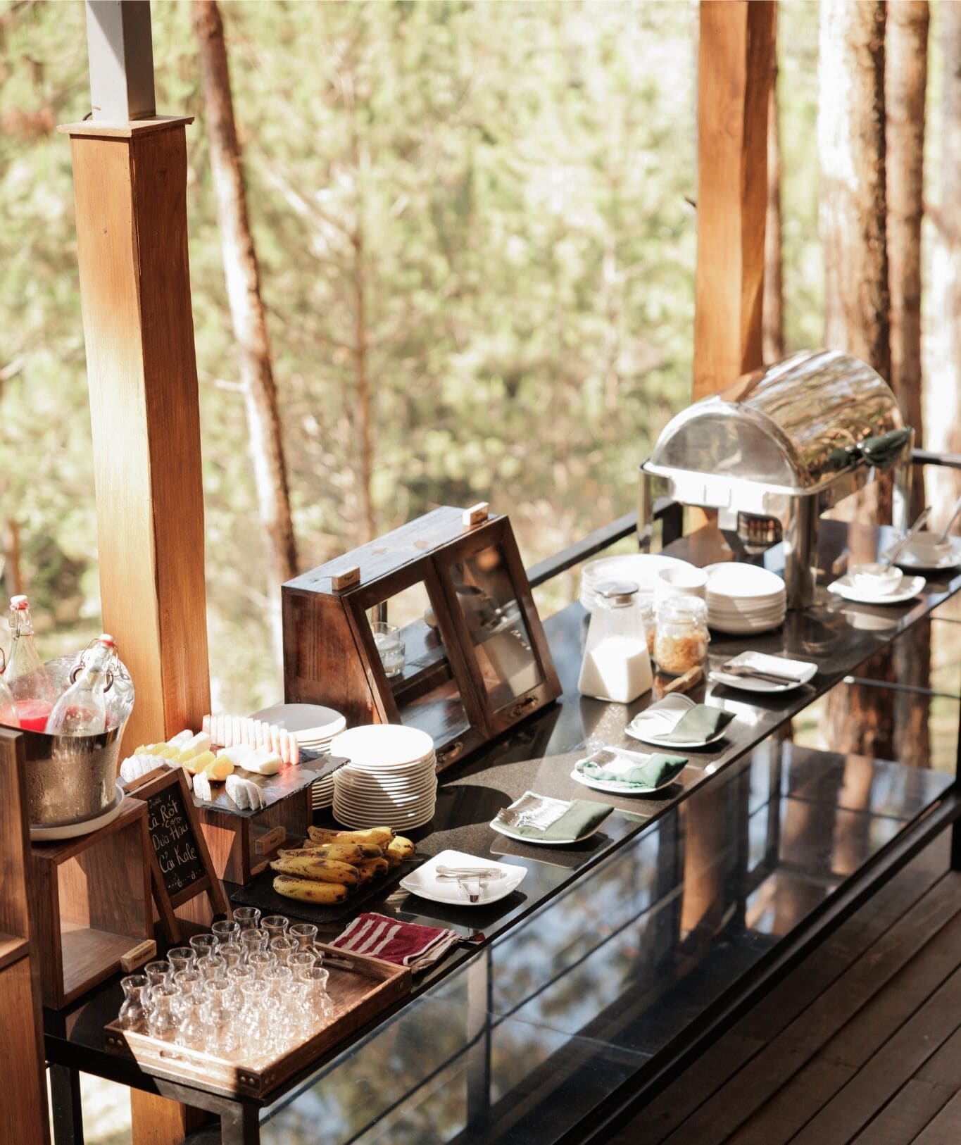 Un buffet de petit-déjeuner est installé sur une table en verre, dans un espace boisé, et propose une variété d’aliments et de boissons en libre-service.