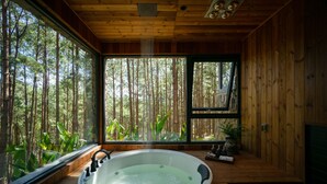 A wooden cabin with a large window, a hot tub, and a view of a forest. - Mơ Stay Forest Resort (Xuan Huong - Da Lat)