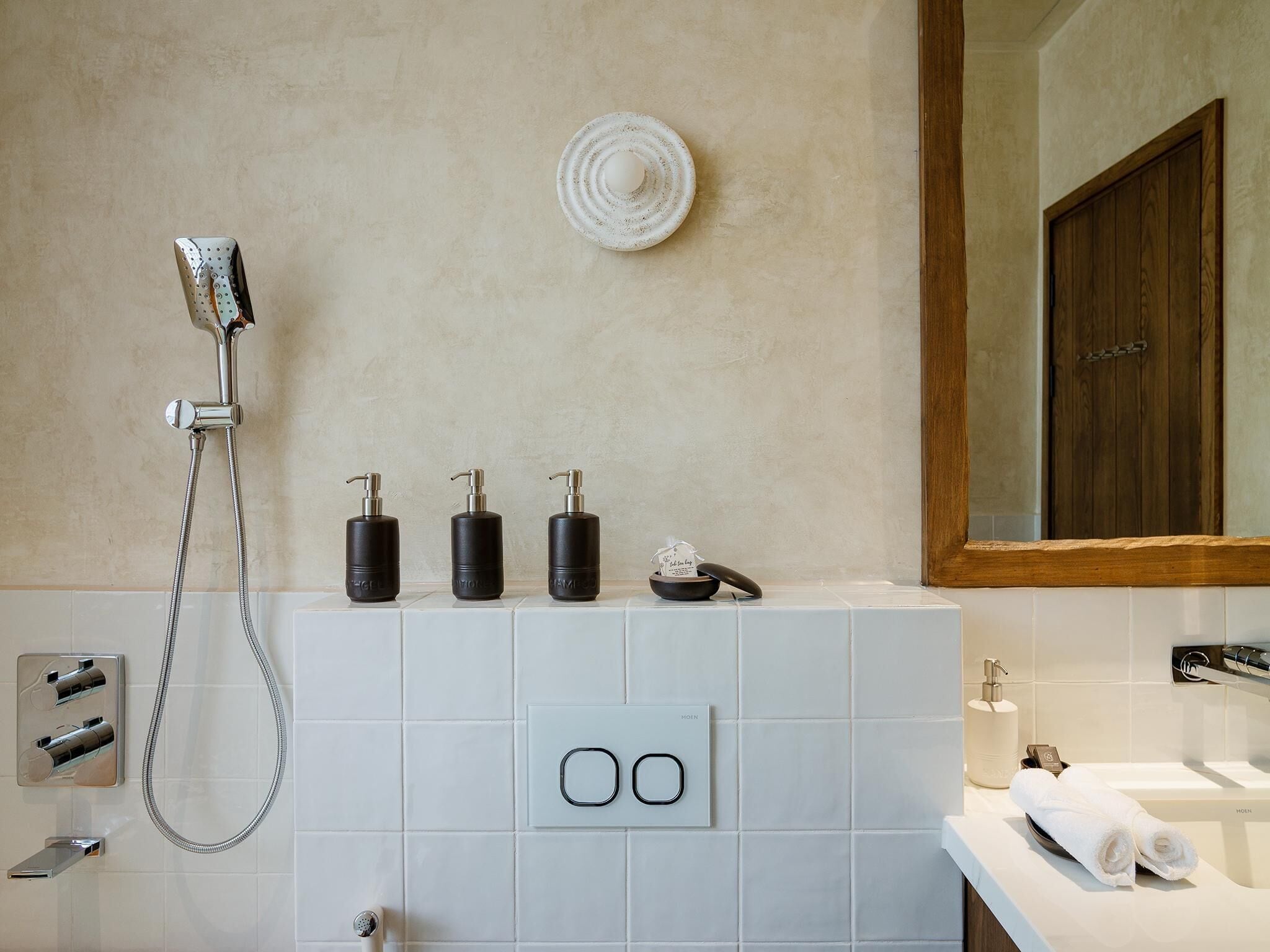 A modern bathroom with a shower, three black soap dispensers, a small bowl, and a towel on the counter.