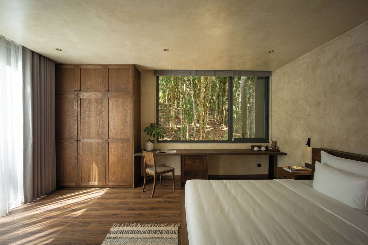 A bedroom with a large window, wooden wardrobe, a desk, and a bed.