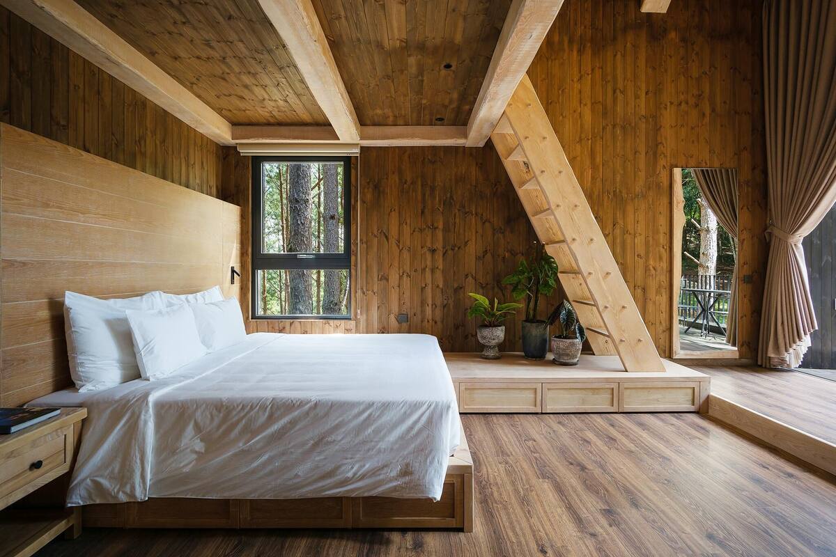 A bedroom with a wooden ceiling, a large bed, a window with a view of trees, and a small seating area with plants.