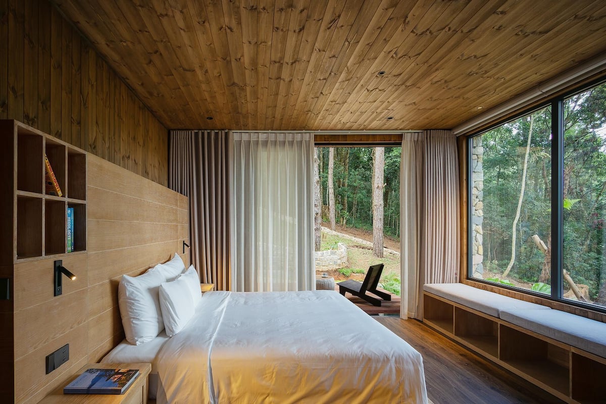 A bedroom with a large bed, wooden headboard, built-in shelving, a bench by the window, and a view of the forest.