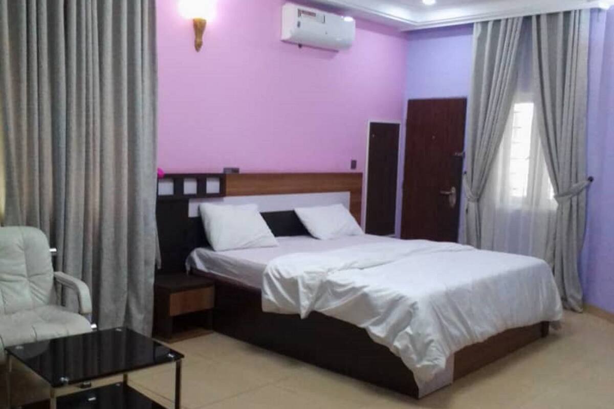 Quarto de hotel com cama, mesa de cabeceira, cadeira, pequena mesa e ar condicionado.