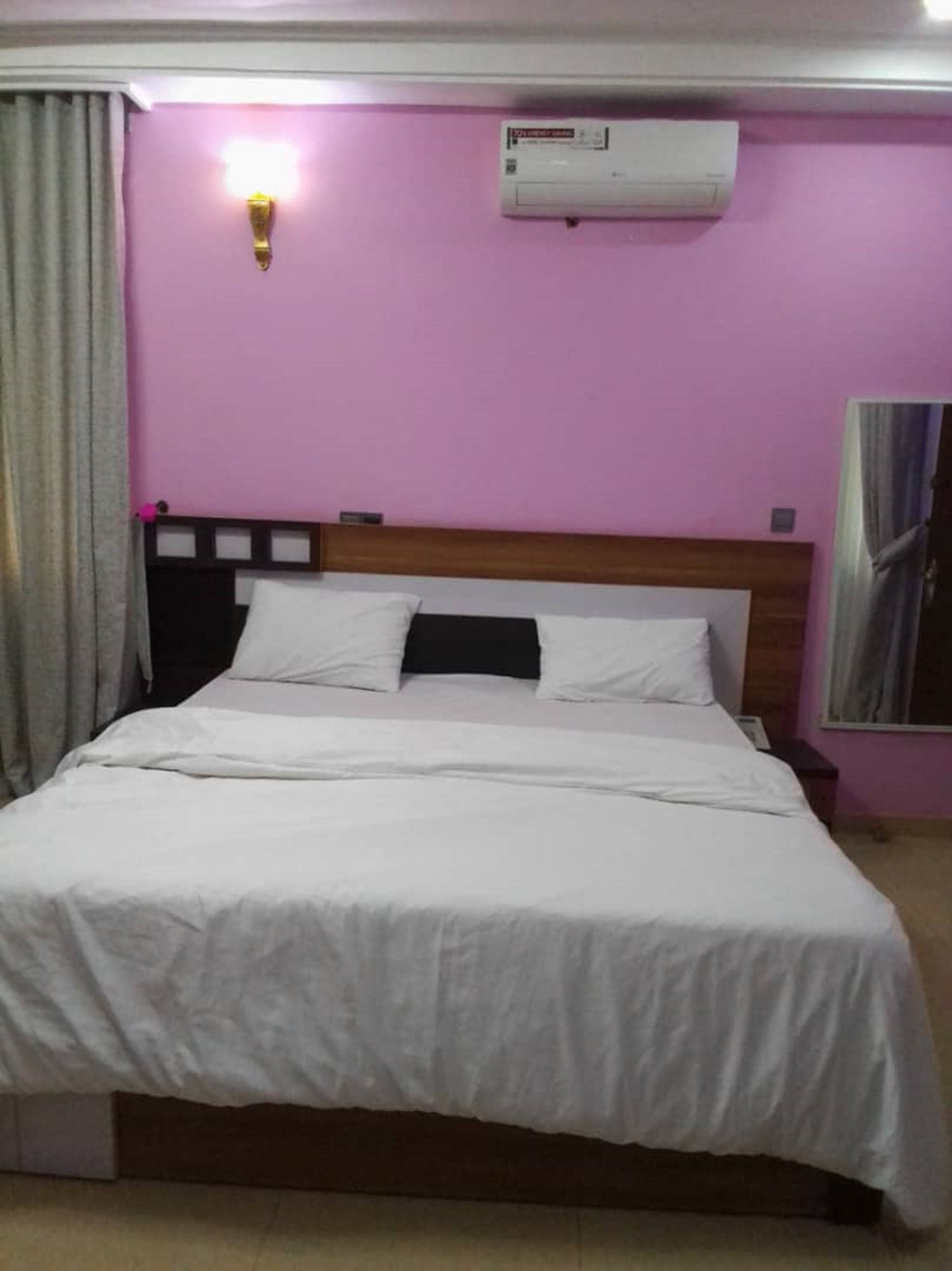 A single bed with white bedding in a room with pink walls, an air conditioning unit above the bed, and a window with curtains to the right.