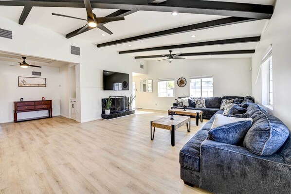 A spacious living room with a large sectional sofa, a coffee table, and a fireplace. There are ceiling fans, a TV mounted on the wall, and a wooden floor.